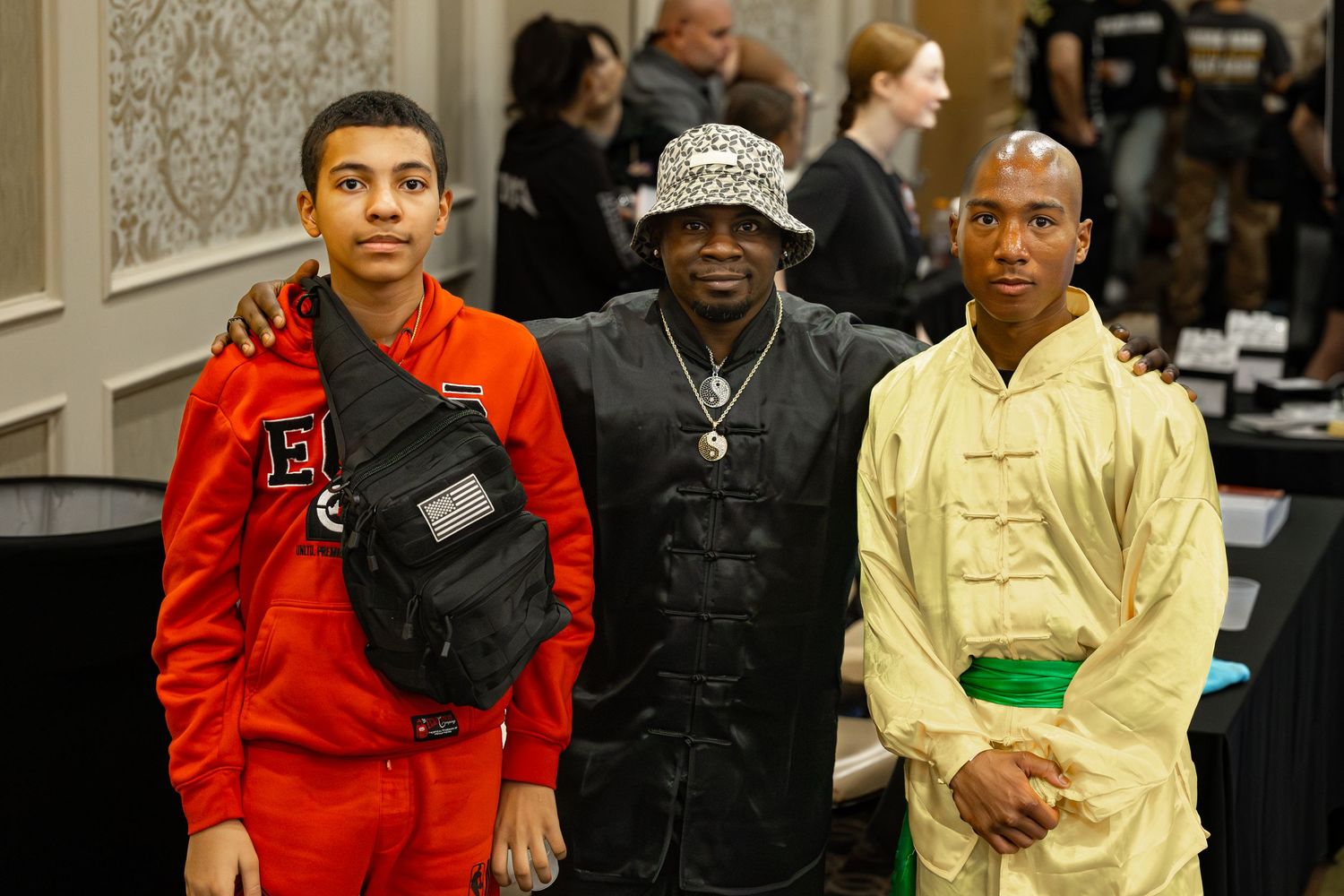 Three men posing together at an indoor event, wearing stylish outfits including red tracksuit, black jacket, and yellow ensemble.