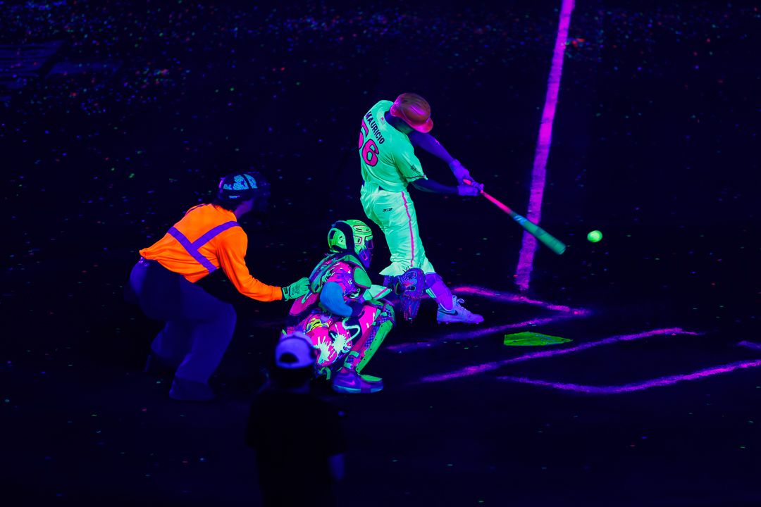 Players in neon UV-reactive costumes play baseball under black light illumination.