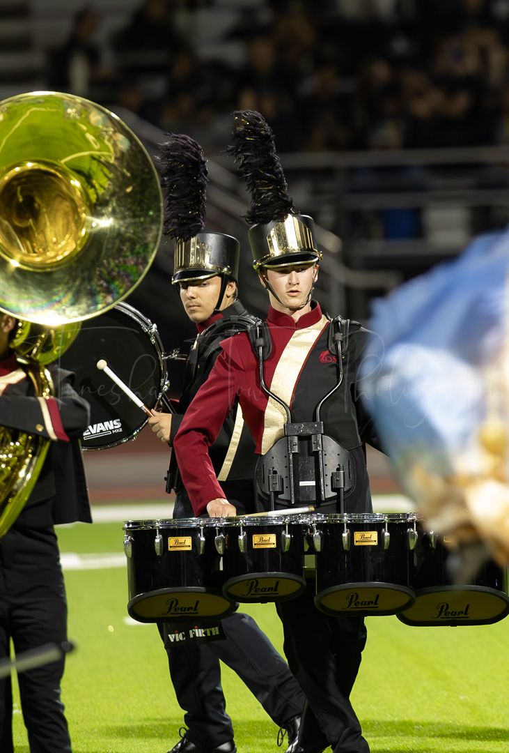 Marching band members in dark uniforms carry brass instruments across a football field.