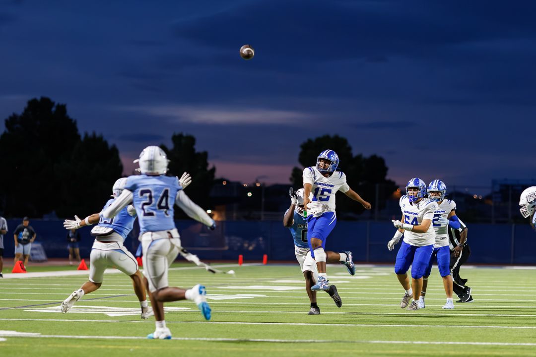 Football players compete under evening sky, with ball mid-air during a high school night game on a well-lit field.