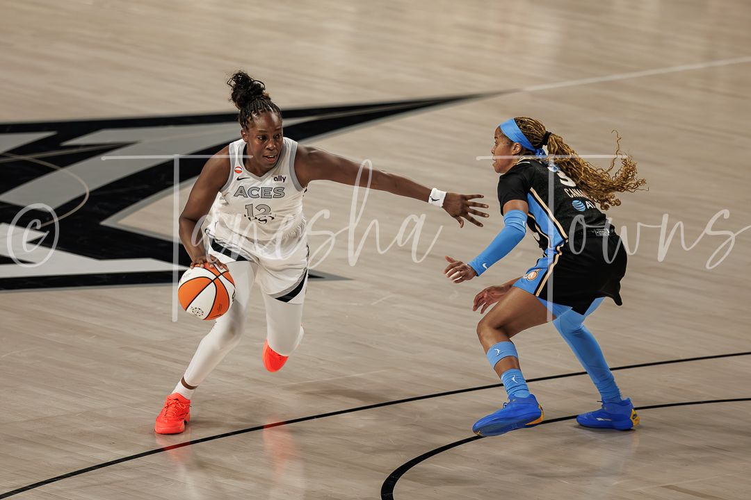 WNBA player in white Las Vegas Aces uniform dribbles past a defender in blue during a professional basketball game.