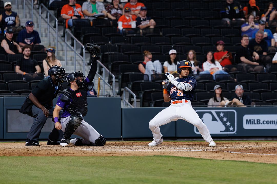 A batter prepares to swing during a MiLB night game as the catcher and umpire watch closely at home plate.
