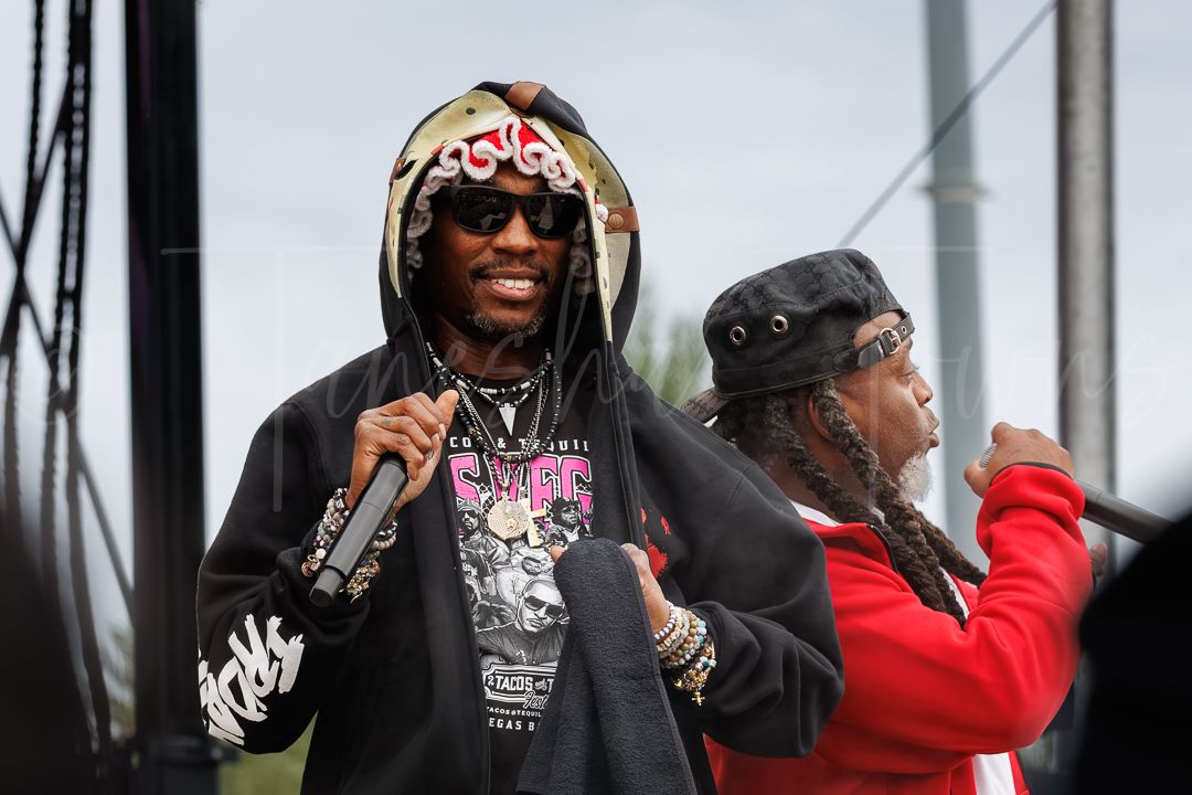 Two male hip-hop artists performing on outdoor festival stage, wearing hoodies and sunglasses while holding microphones.