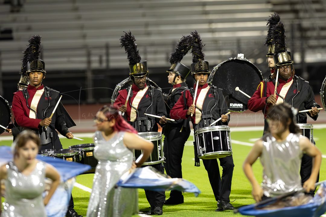Marching band performers in black and red uniforms perform on a football field at night.