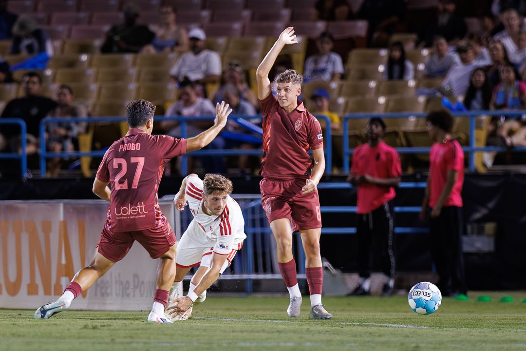 College soccer players in maroon and white uniforms compete for the ball during a night game on a well-lit field.