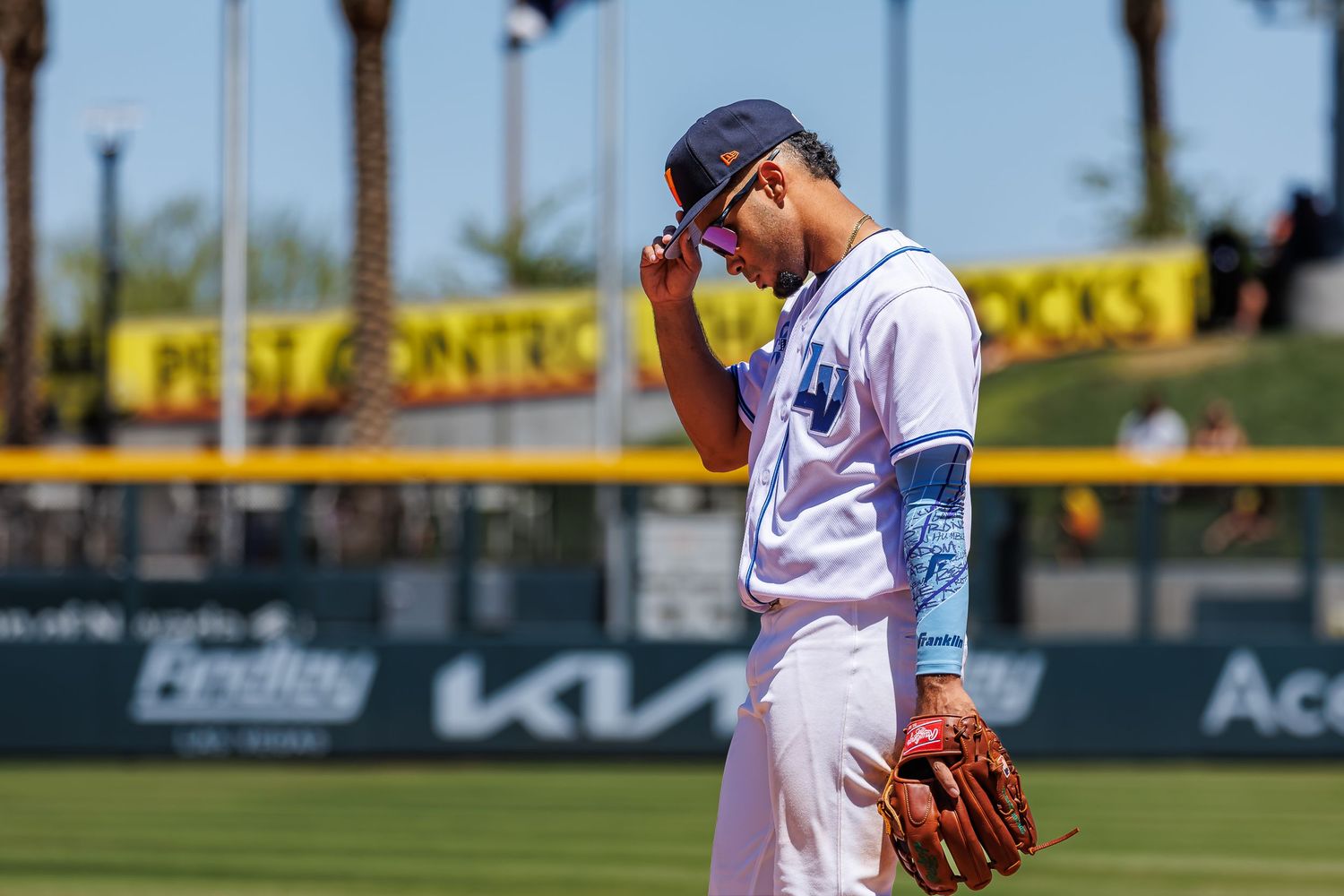 Baseball player in blue uniform adjusts cap on sunny field during a game.