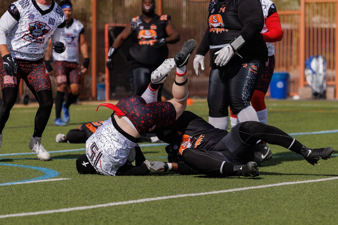 American football players tackling during an outdoor game on a green turf field, wearing black and orange uniforms.