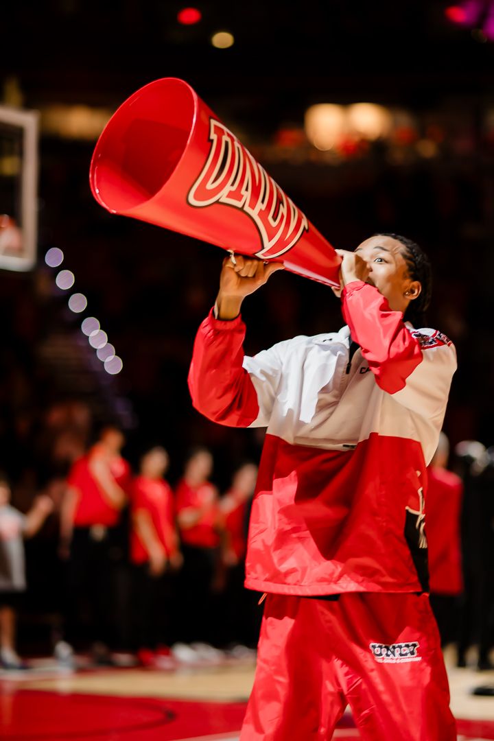 Cheerleader in red uniform performs energetic routine with megaphone during indoor sporting event.