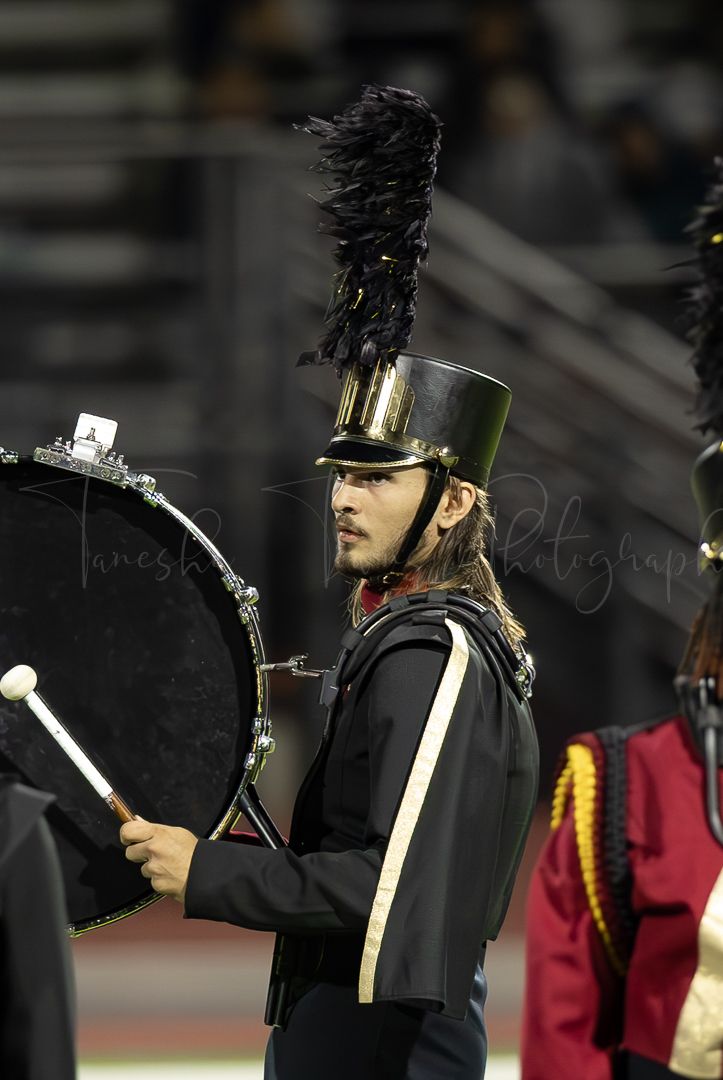 Marching band member in black uniform and tall hat performing at night.