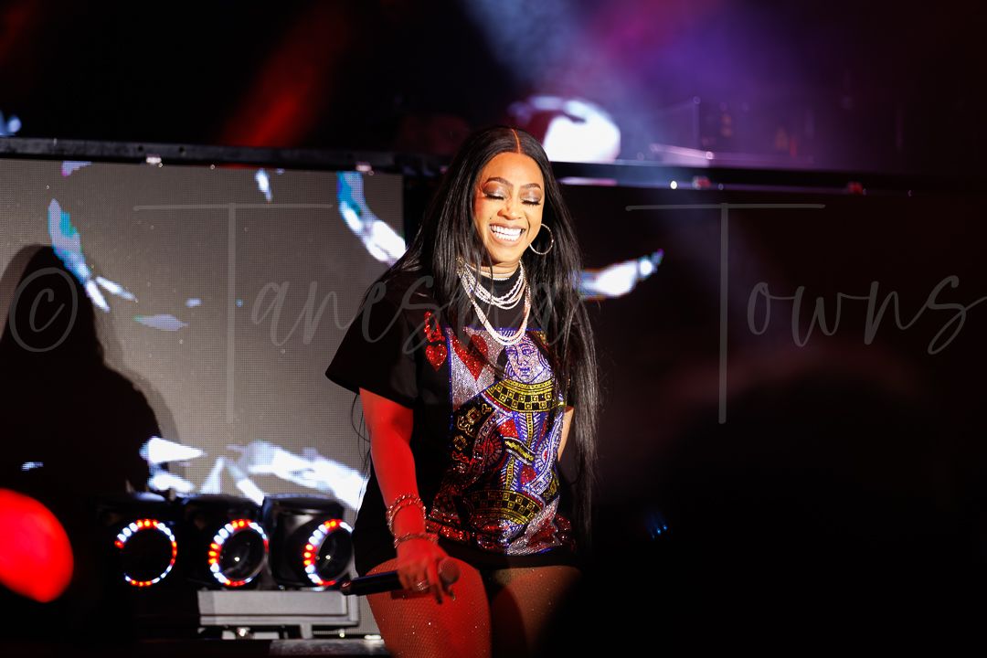 Female performer on stage in colorful graphic tee and layered necklaces, smiling under dramatic concert lighting.