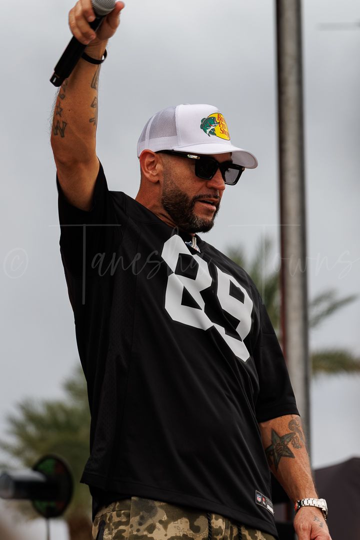 Man in black jersey number 29, white trucker hat, and sunglasses holds microphone at outdoor event.