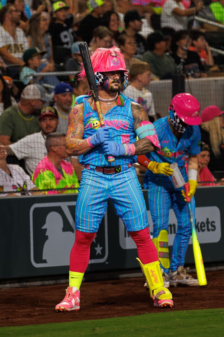 Baseball player in vibrant pink and blue neon costume holding bat at MLB celebrity game event.