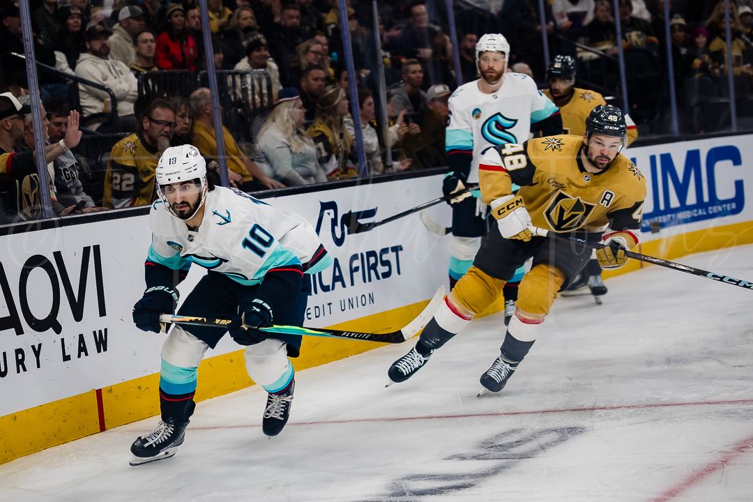 NHL players from the Sharks and Golden Knights chase after the puck along the boards.