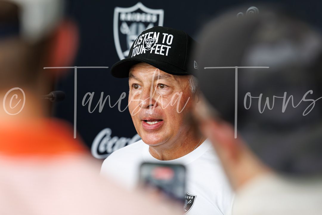 Raiders coach speaks to media at training camp wearing a Raiders hat and white shirt.