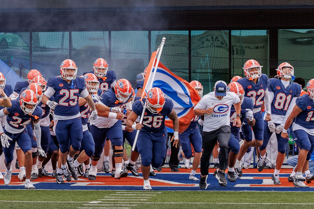 College football team in blue and orange uniforms charging onto field with flag bearer leading the charge.