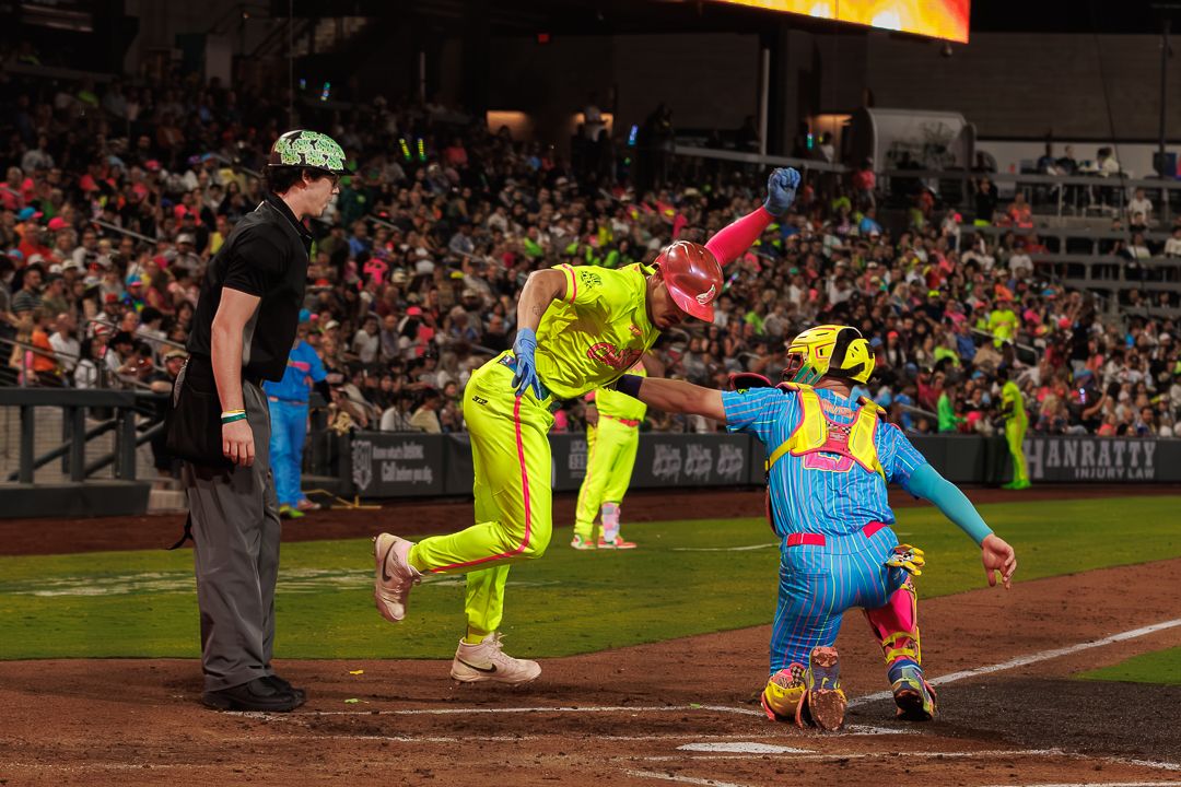 Colorful baseball players celebrate at home plate during a vibrant night game under bright stadium lights.