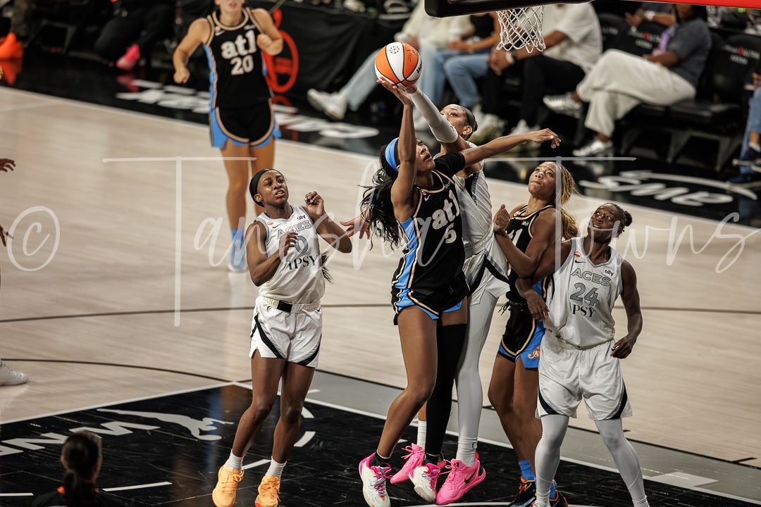 WNBA players compete for a rebound during an intense basketball game, with one player in black driving to the basket.