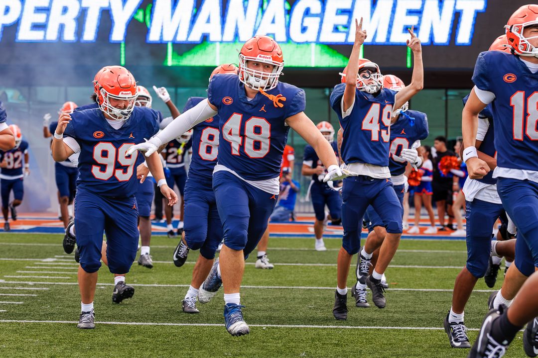 College football players in blue uniforms numbered 99, 48, and 49 run onto field through smoke during pregame.