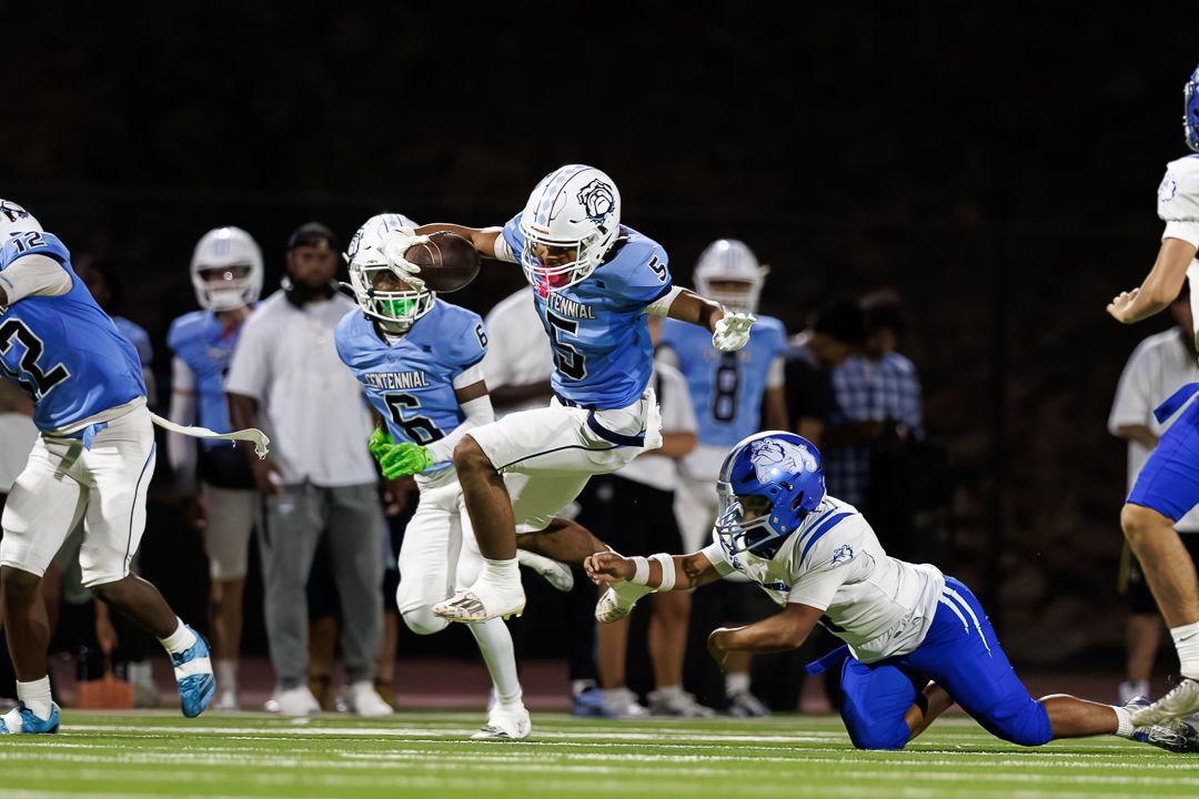 Football players in light blue uniforms compete during a night game, with one player leaping over another during a play.