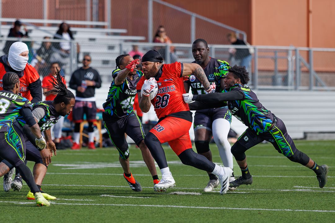 Football player in red jersey #28 breaks through defenders in green during an outdoor game on a turf field.