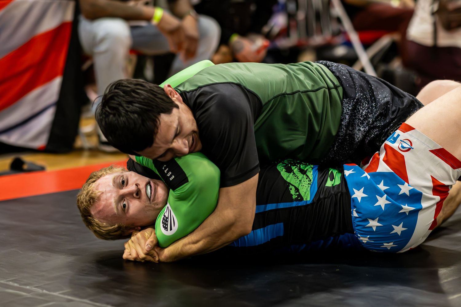 Two wrestlers compete on a mat, with one athlete in a black and green rash guard applying a submission hold.