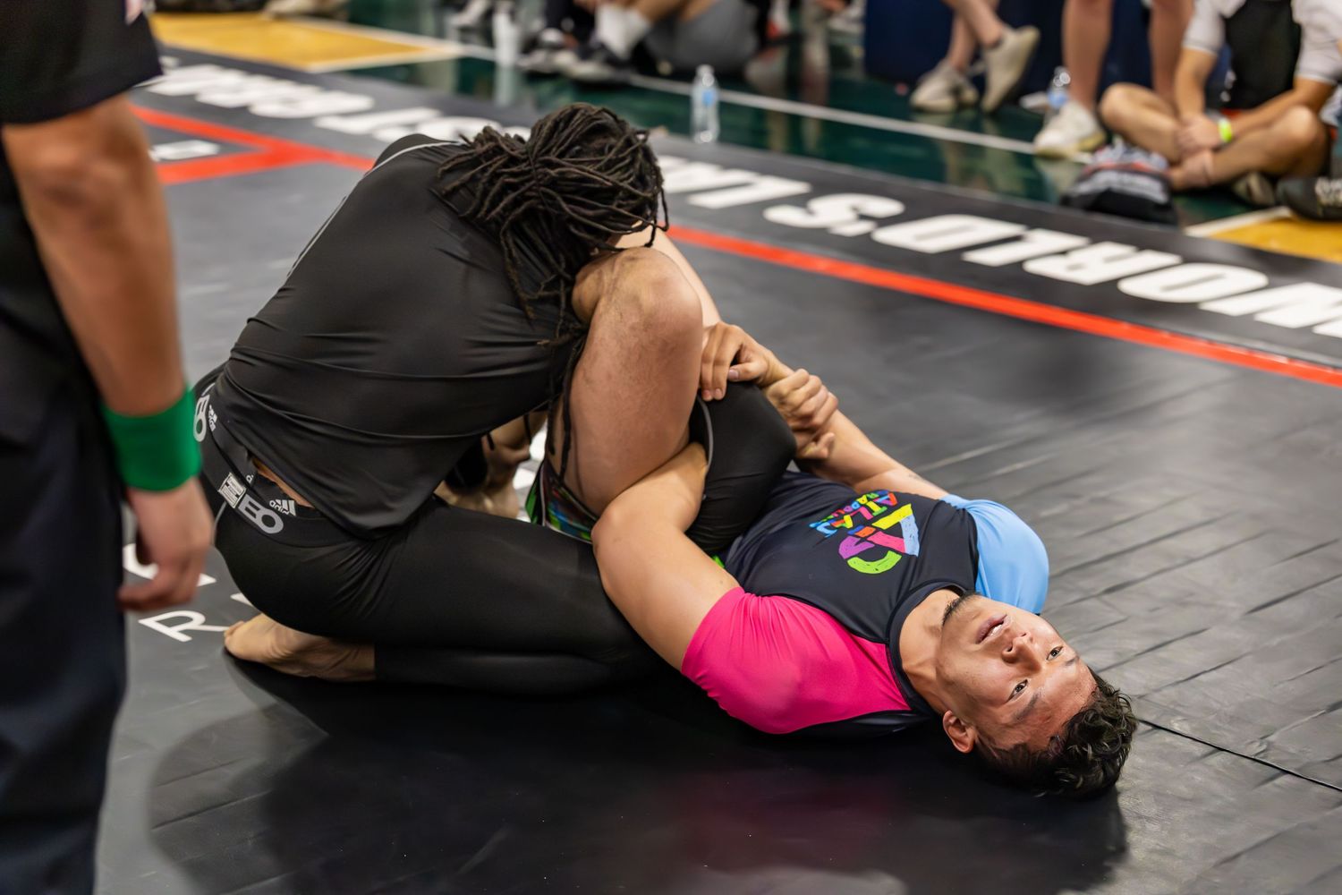 Two competitors grappling on the mat during a Brazilian jiu-jitsu tournament match.