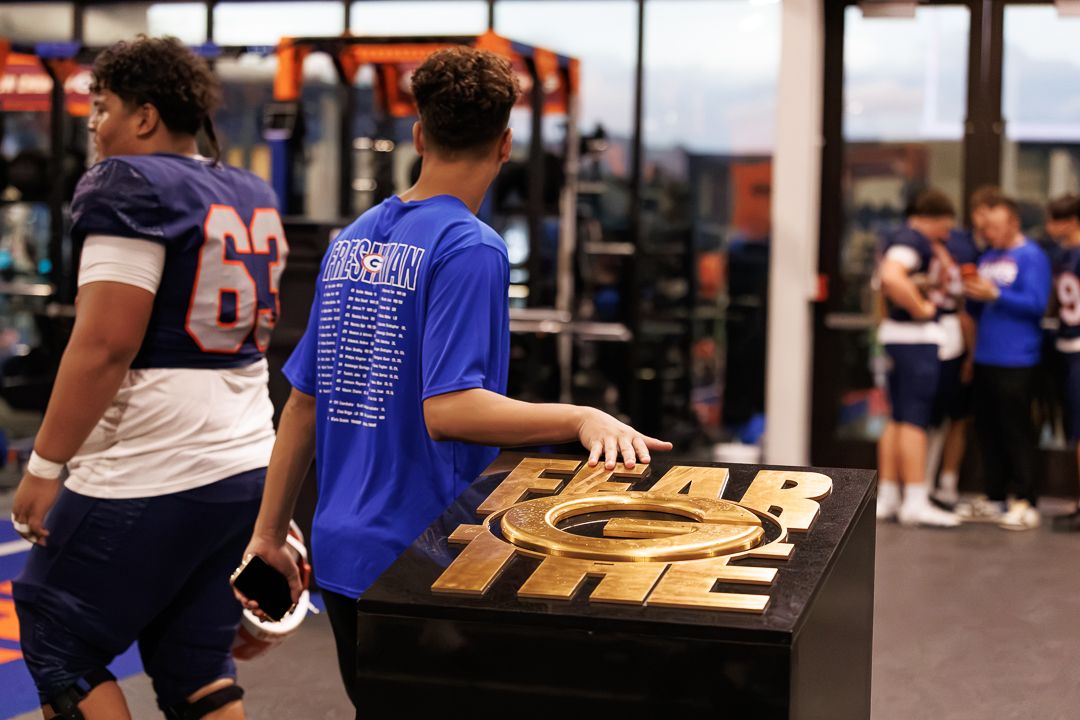 Football player in blue jersey reads a trophy display in a modern sports training facility.