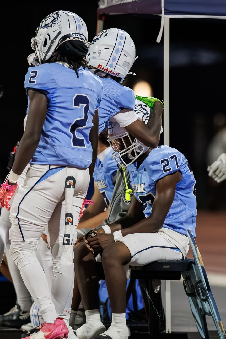 High school football players in light blue uniforms resting on the sideline bench during a night game.