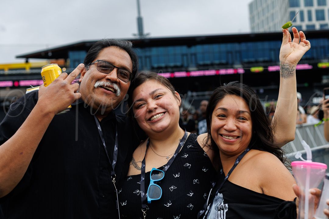 Three friends smile and pose together at an outdoor music festival, holding drinks and enjoying the event.
