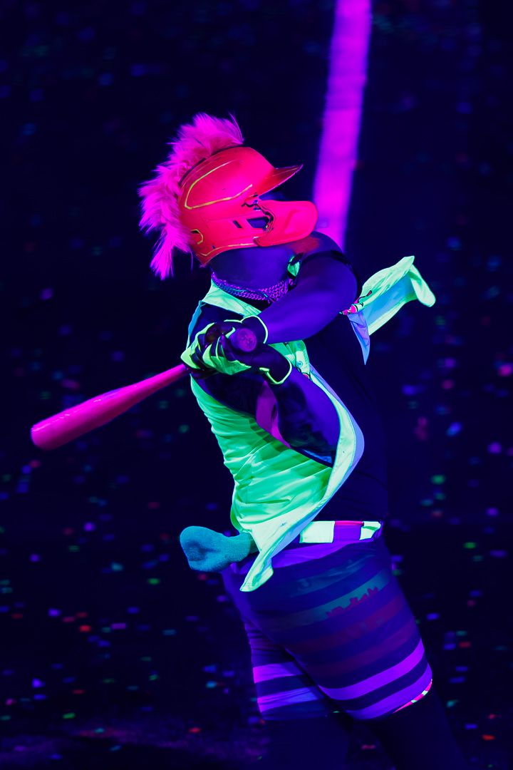 Performer in vibrant neon UV outfit swings a baseball bat under black light.