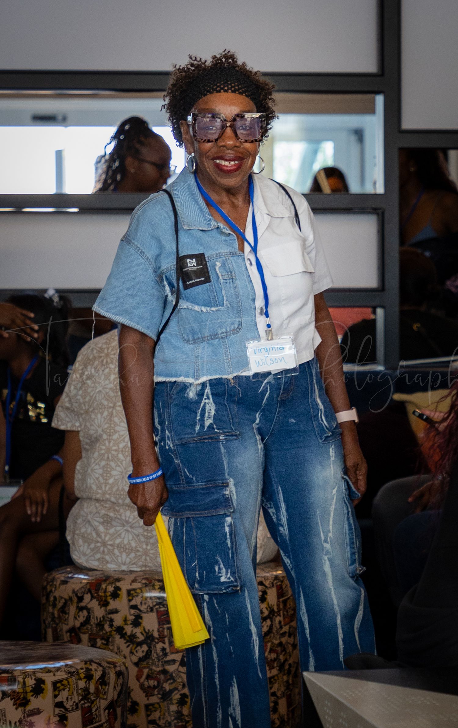 Participant in denim outfit and glasses poses during indoor gathering.