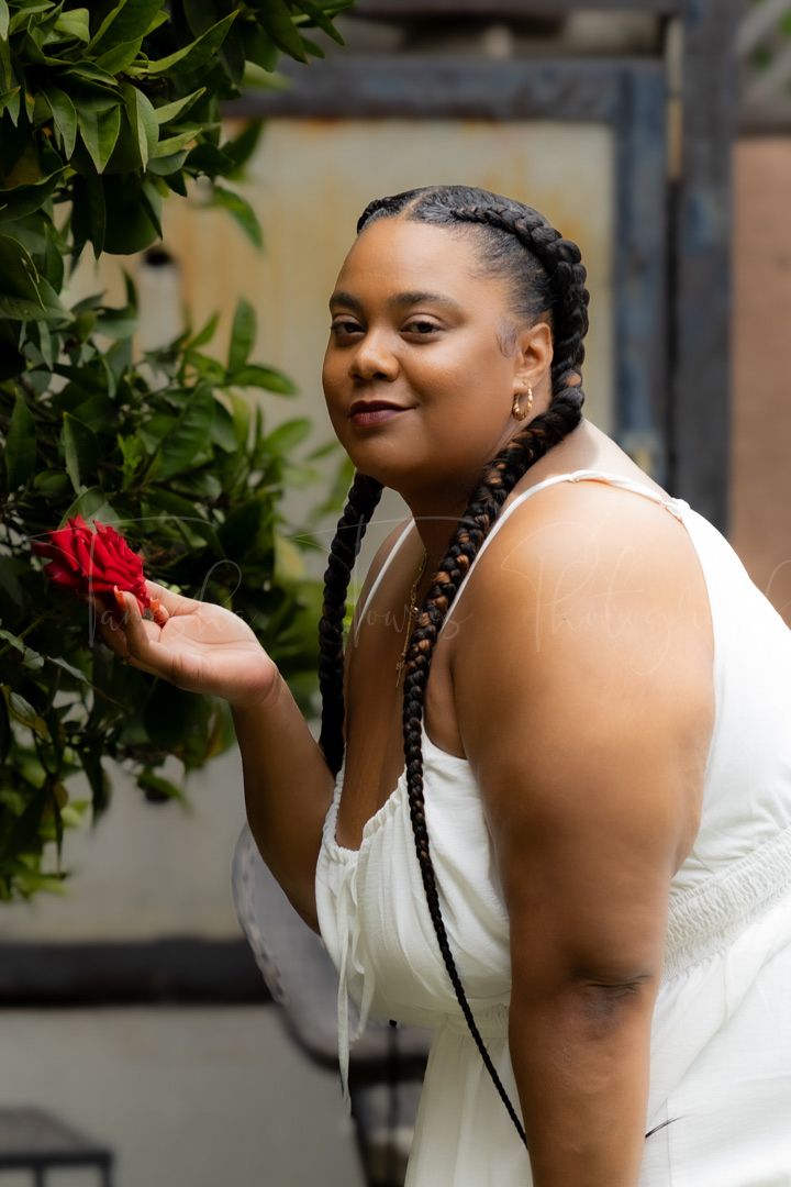 A person in a white dress poses with a red flower in a garden setting.