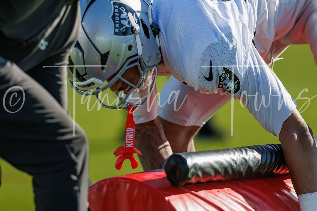 Football player in white jersey and helmet drives into a blocking pad during an intense practice drill on a sunny field.