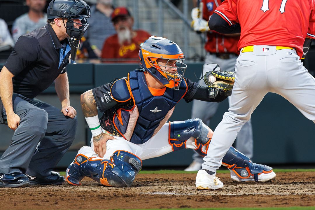 Baseball catcher in orange helmet crouches to receive pitch as batter swings during a game.