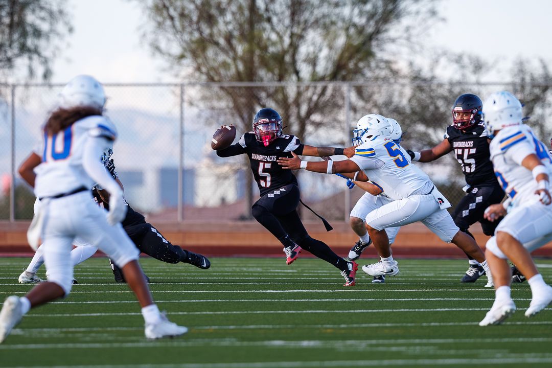 Football player #5 in red uniform extends for a catch while blue team defenders attempt to make a tackle on the field.