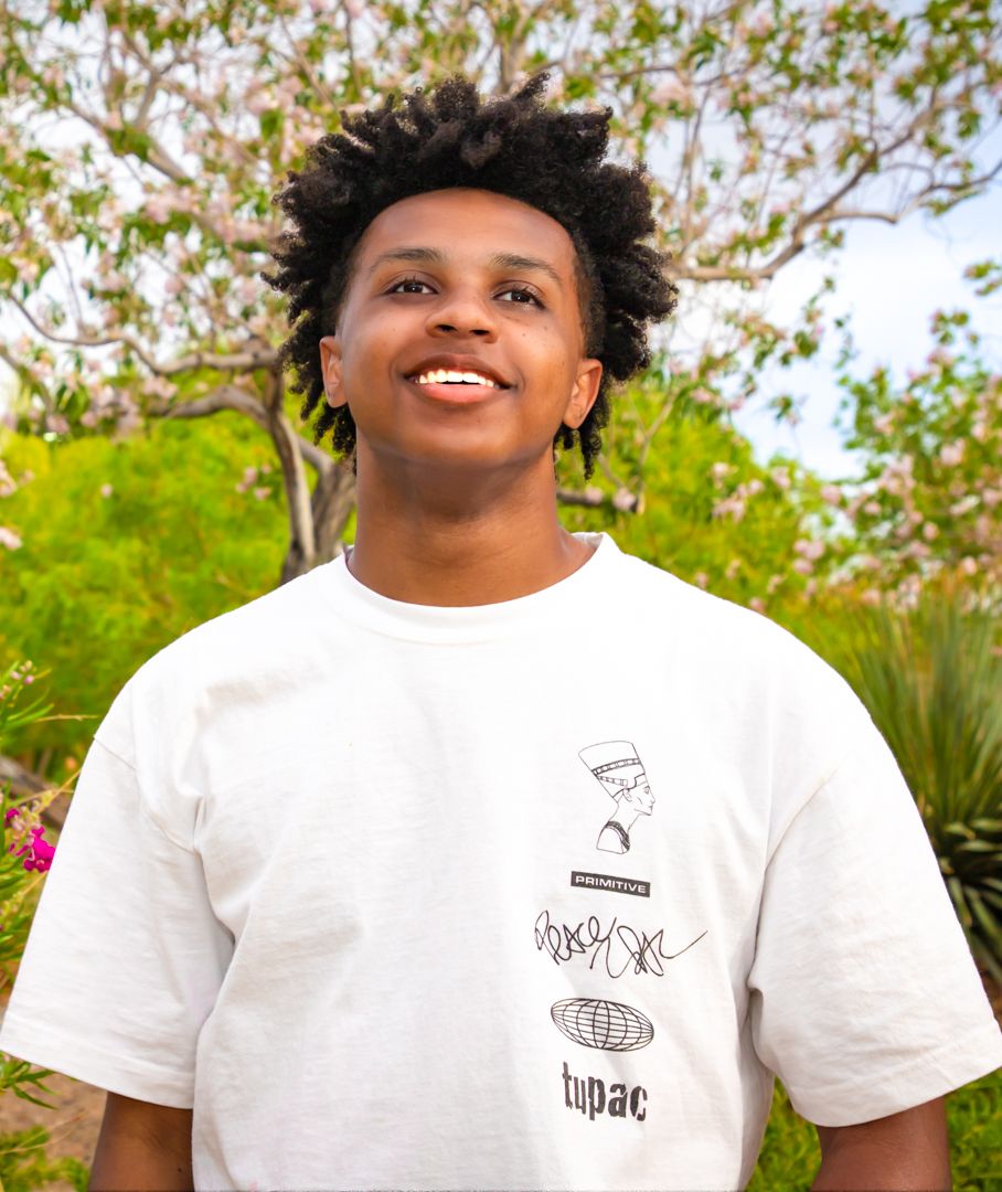 Person in white t-shirt smiles while standing in a garden setting with trees and plants.