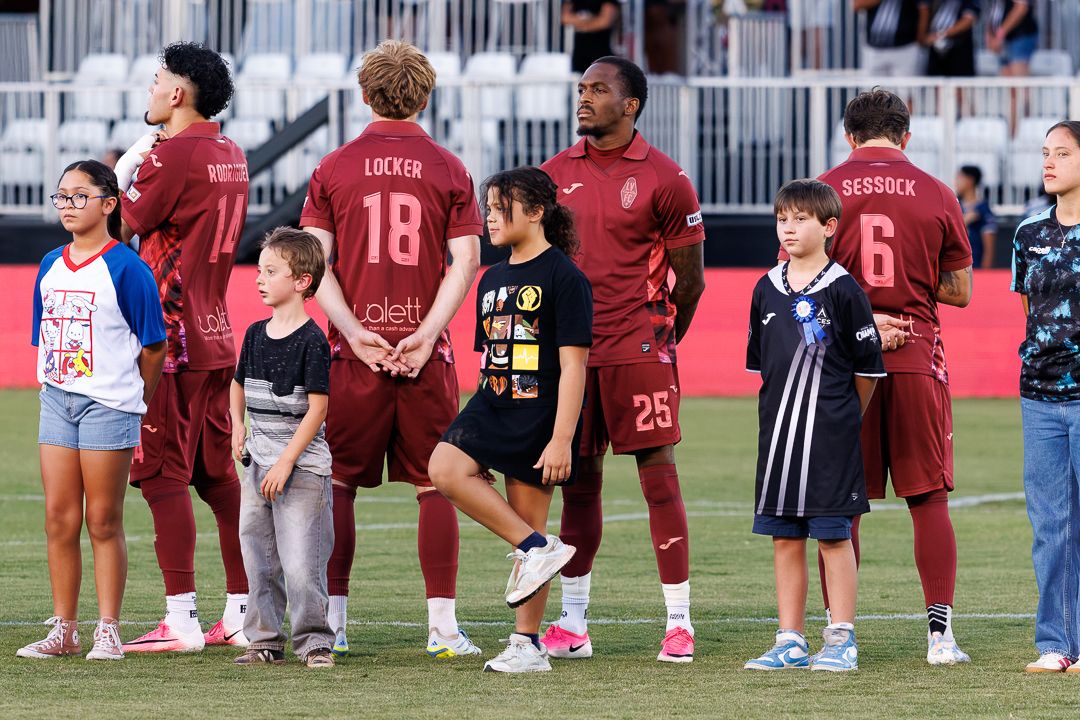 Soccer players in red uniforms with numbers 18, 25, and 6 stand with young fans on a field before a match.