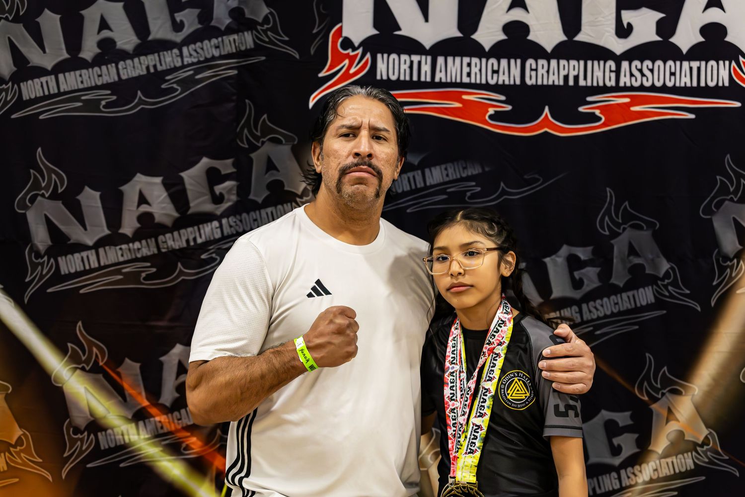 Two competitors pose proudly at a NAGA North American Grappling Association tournament, with medals displayed.