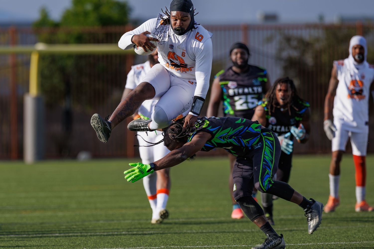 Athletes compete in a flag football game, with one player leaping over a diving defender on a green field.