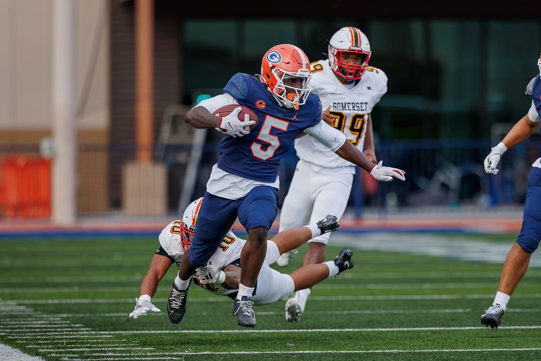Football player #5 in blue uniform runs with the ball while being pursued by defenders in white during a college game.