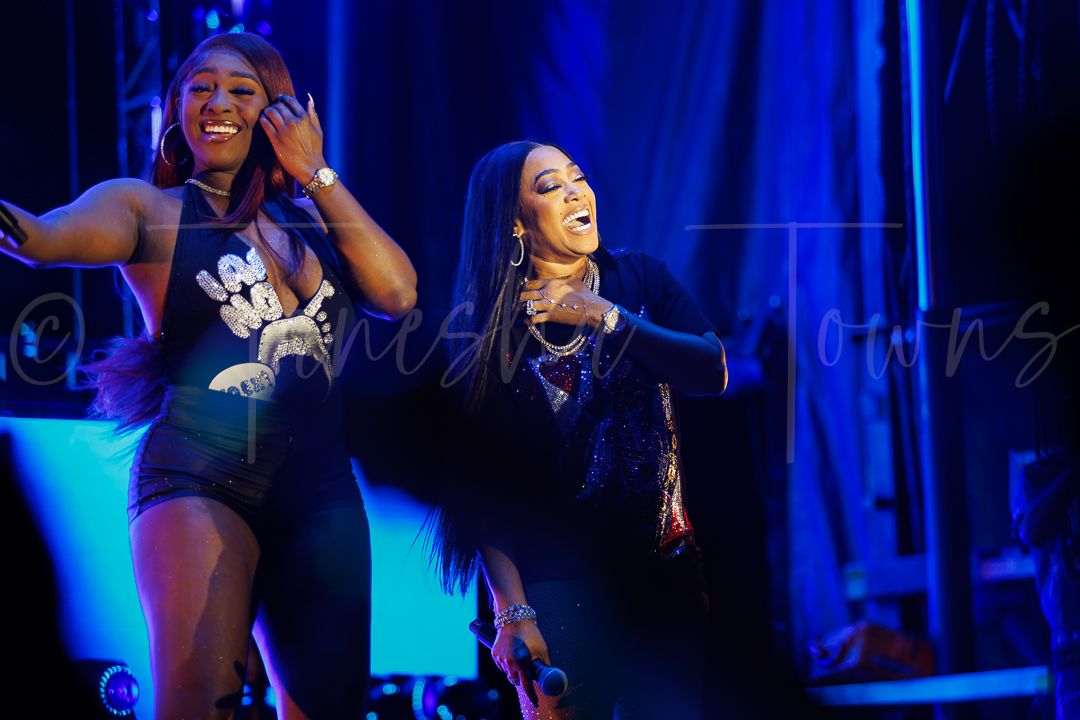 Two women laughing and dancing joyfully together on a blue-lit concert stage.