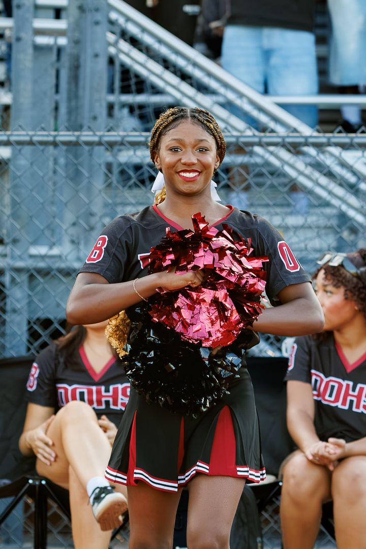 Smiling cheerleader in black and red DOHS uniform holds red and black pom-poms at a football game.