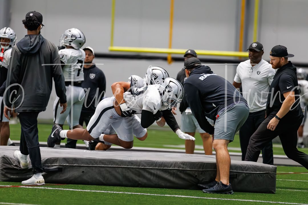 NFL players engage in blocking drills over a practice pad during an indoor Raiders training camp session.