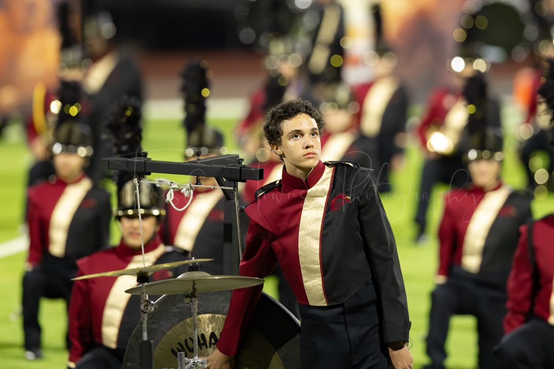 Marching band members in red and black uniforms perform with colorful field lighting.