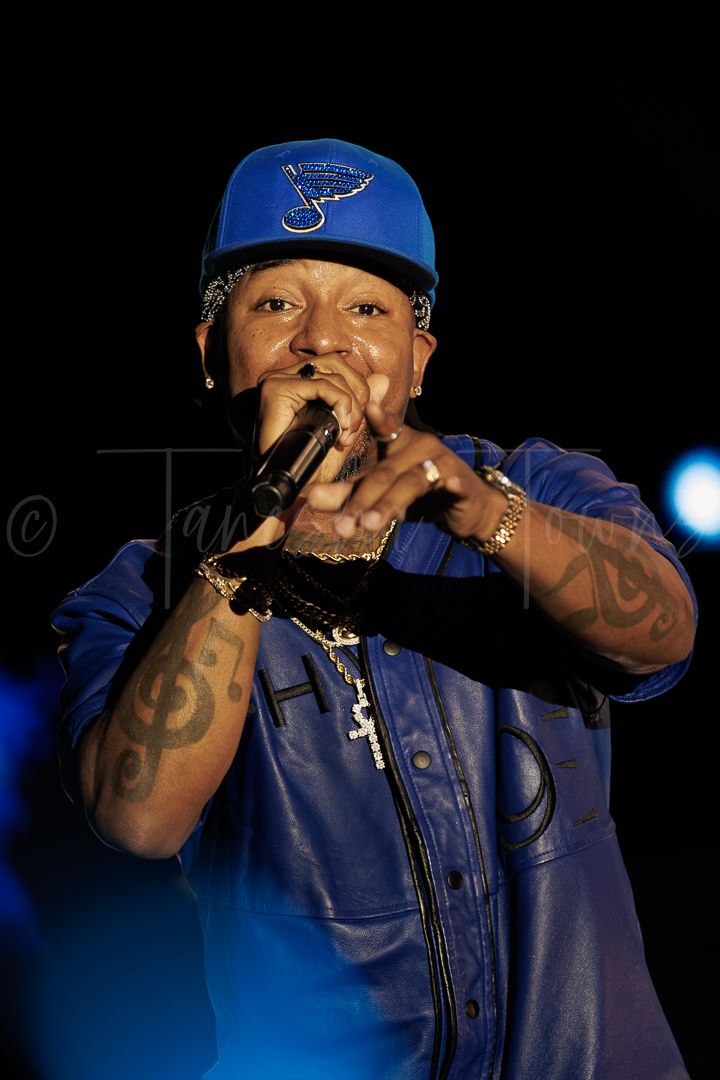 Male rapper performing on stage wearing blue cap and jacket, pointing at crowd while holding microphone.