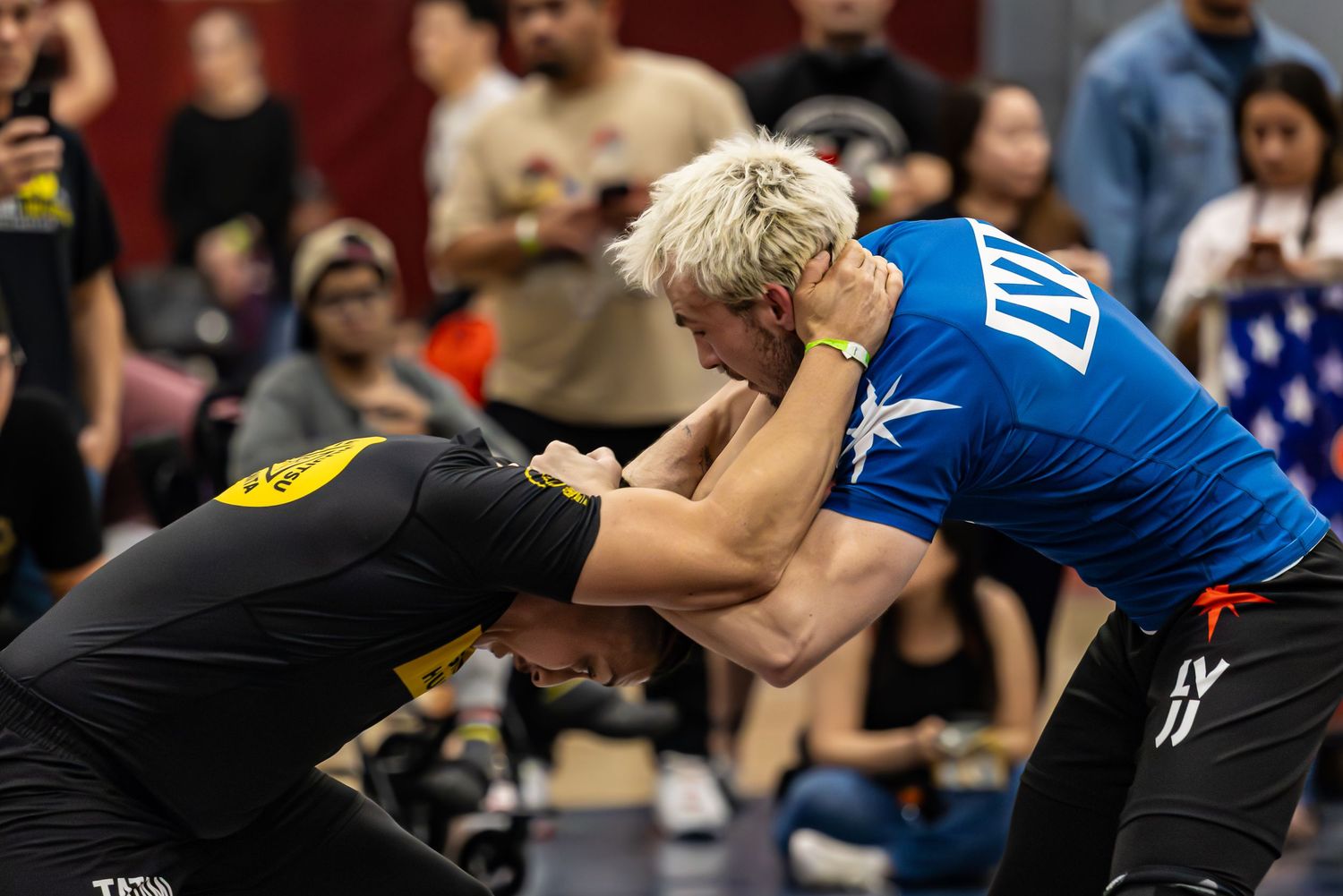 Two athletes compete in a grappling match at a martial arts tournament, with spectators watching in the background.