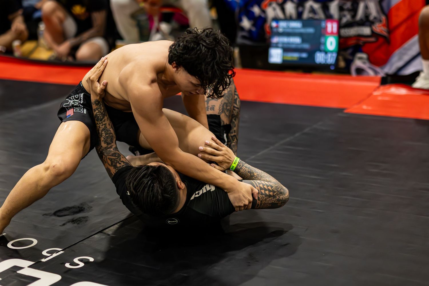 Two tattooed MMA fighters grappling on a black mat during a submission wrestling competition.