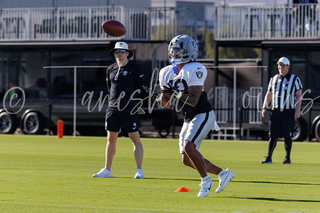 NFL player catches a football during a Raiders practice session on a green field with coaches and officials watching.