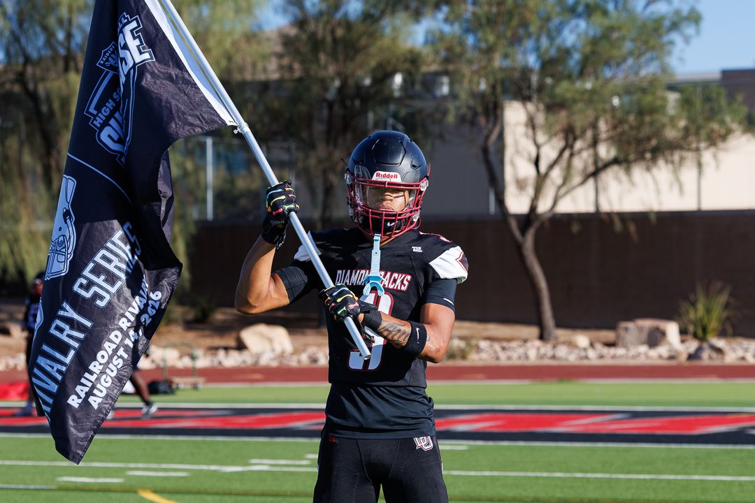 Football player in black uniform holds team flag on red turf field during game day.