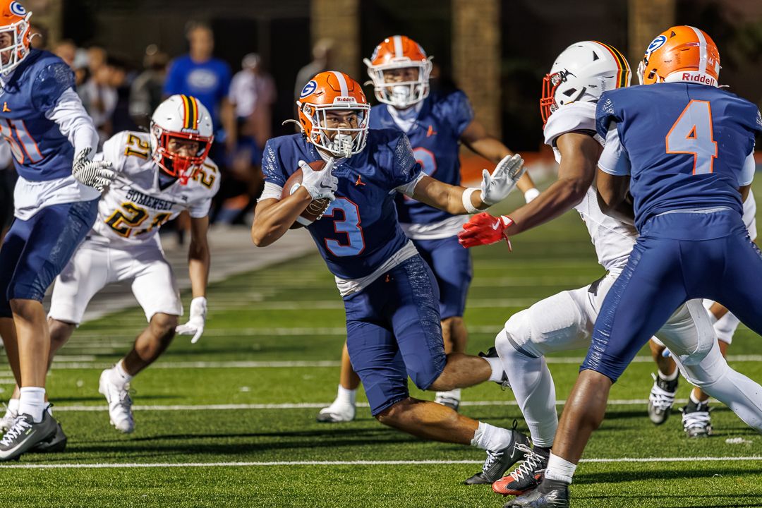 Football player #3 in blue uniform runs with the ball during a night game, evading defenders on the field.